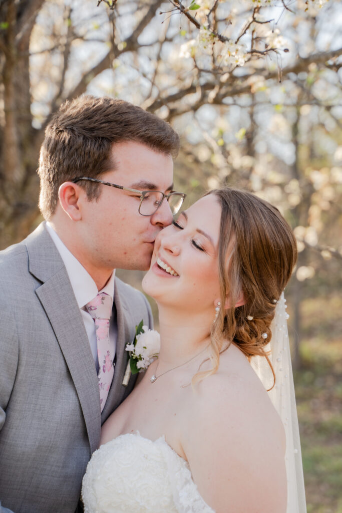 groom kissing bride on the cheek with a white tree in the background